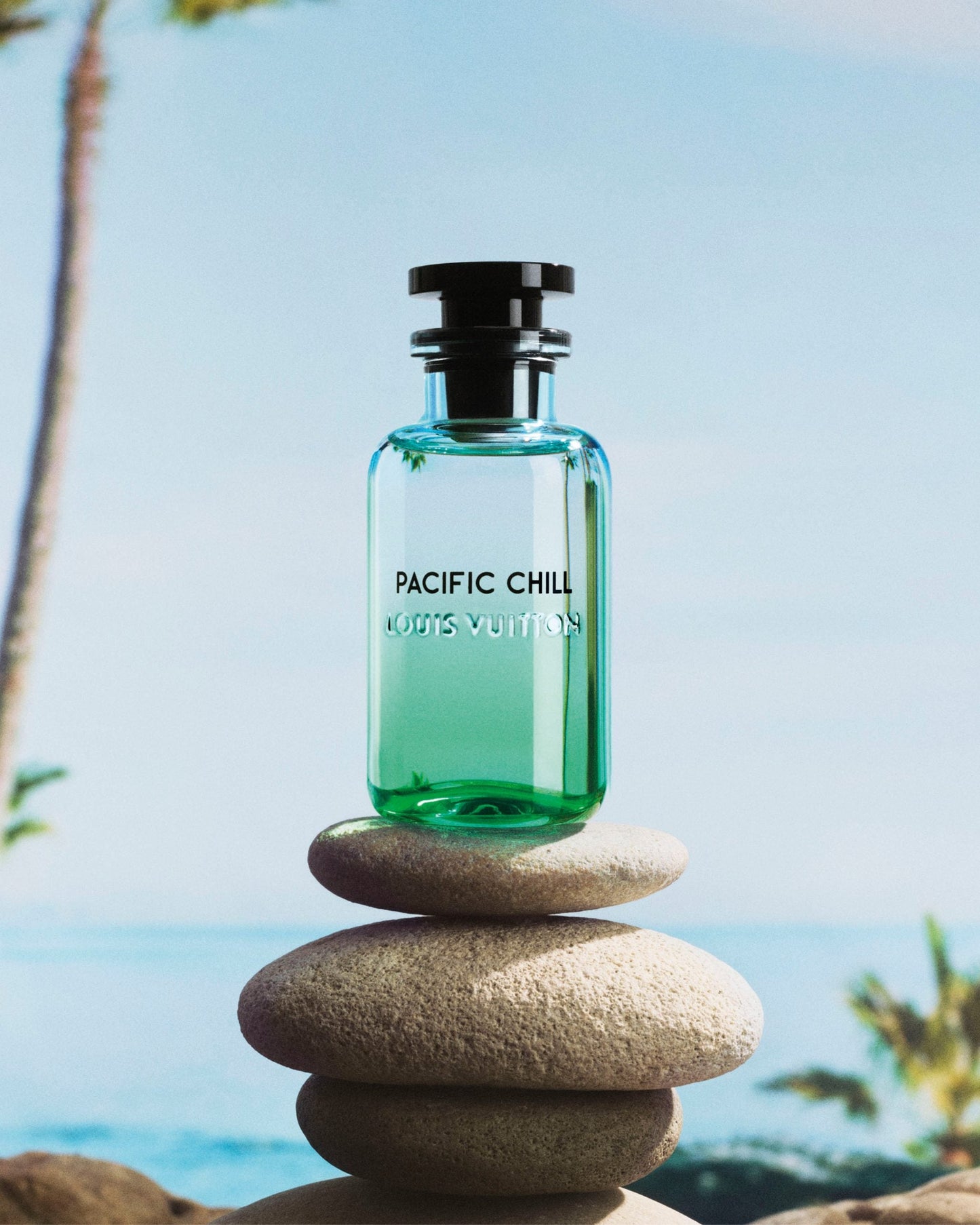 Green perfume bottle labeled 'Pacific Chill' by Louis Vuitton on stacked stones with a beach background.