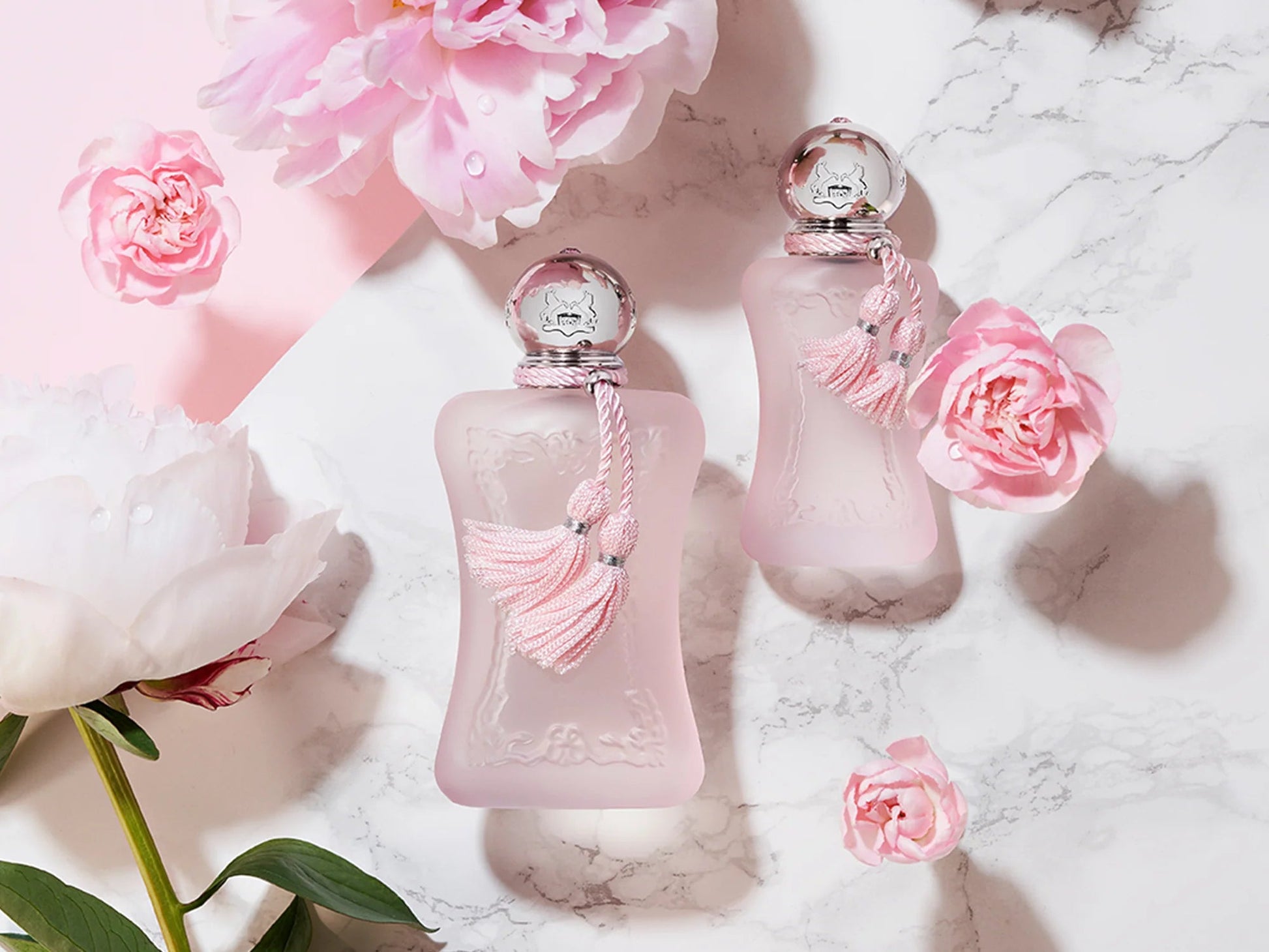 Two pink perfume bottles with decorative elements on a marble surface with flowers.